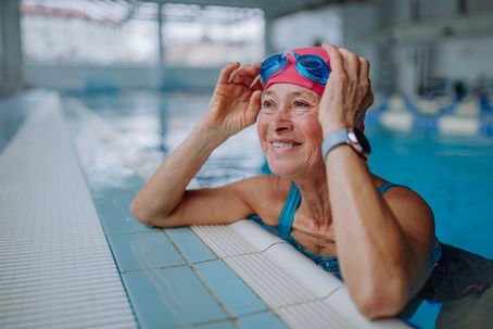 Woman in a pool, smiling, wearing a pink swim cap and blue goggles. Indoor setting with tiled edge and blurred background.