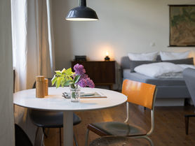 Runder weißer Esstisch mit Stuhl und Salz & Pfeffer und Blumen. Darüber hängt eine Lampe und im Hintergrund sieht man Couch, Bett und Nachttisch sowie eine Landkarte an der Wand.