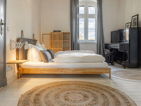 Schlafzimmer mit Nachttischlampe, kleinem Kleiderschrank und Klavier mit Bildern oben drauf. Vor den Betten liegen zwei runde Juteteppiche.