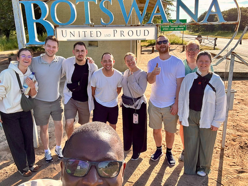 Teilnehmende stehen lächelnd vor dem „BOTSWANA – United and Proud“-Schild an einer Straße nahe der Kasane Transit Route, aufgenommen als Gruppen-Selfie in der Abendsonne.