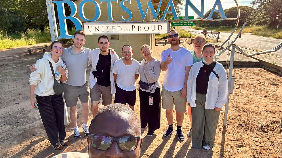 Teilnehmende stehen lächelnd vor dem „BOTSWANA – United and Proud“-Schild an einer Straße nahe der Kasane Transit Route, aufgenommen als Gruppen-Selfie in der Abendsonne.