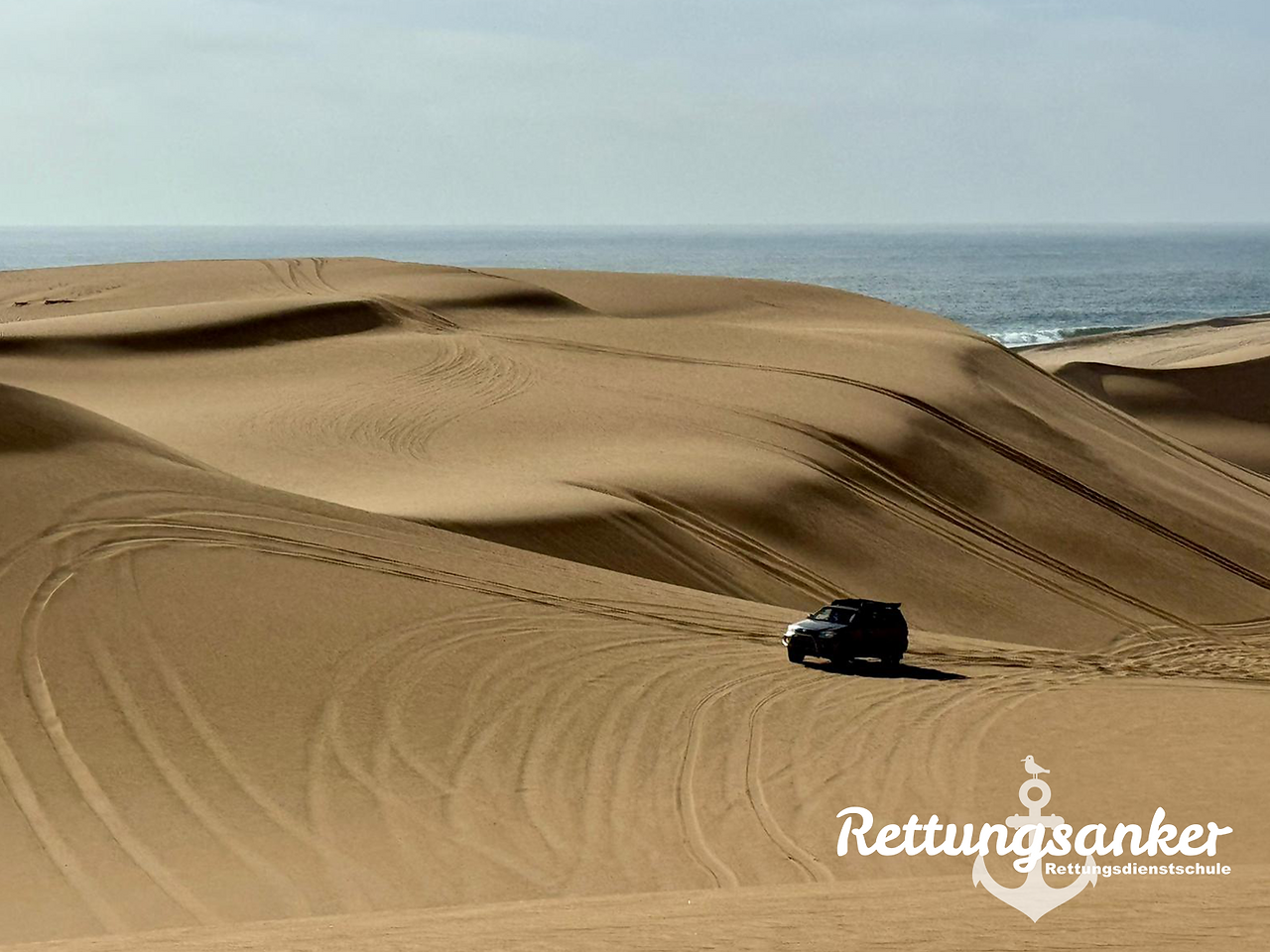 Geländewagen fährt durch die Dünen bei Walvis Bay; im Hintergrund sind Atlantik und Küstenlinie zu sehen.