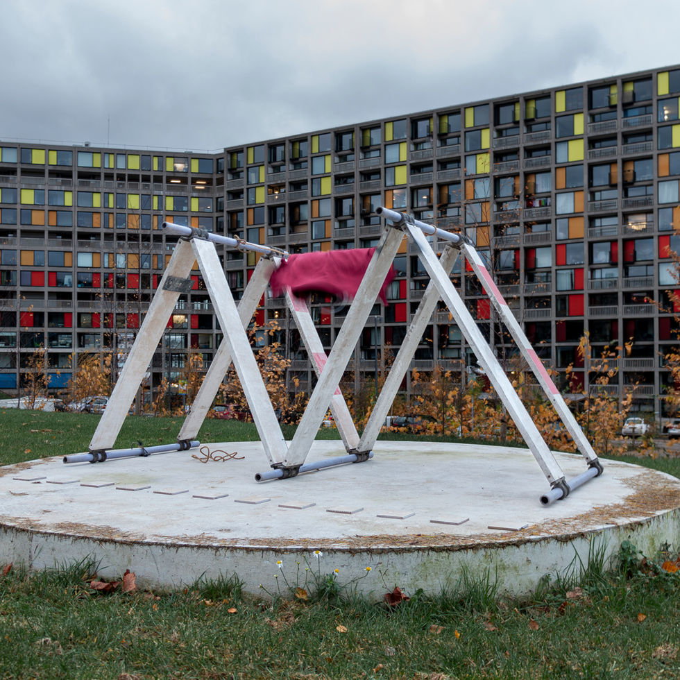 ‘Back-to-back (Obstructions) artwork by artist Tyler Mallison, featuring a deep red textile t-shirt suspended between two thin triangular forms and small square tiles set out on a large round concrete plinth. The modern, abstract installation is in a distinctive architectural setting within  the grounds of the large listed ‘Park Hill Estate’ residential complex with brightly coloured panels in view. 