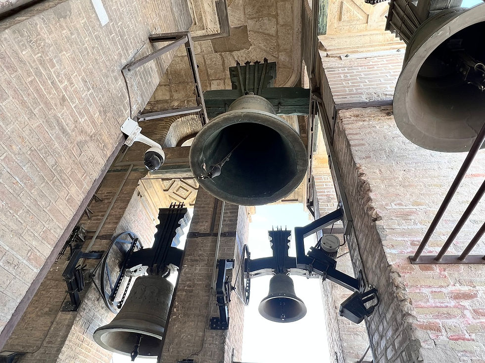 Dzwony kościelne zawieszone na metalowych wspornikach w murowanej wieży. Jasne cegły w tle tworzą surowe, historyczne otoczenie.