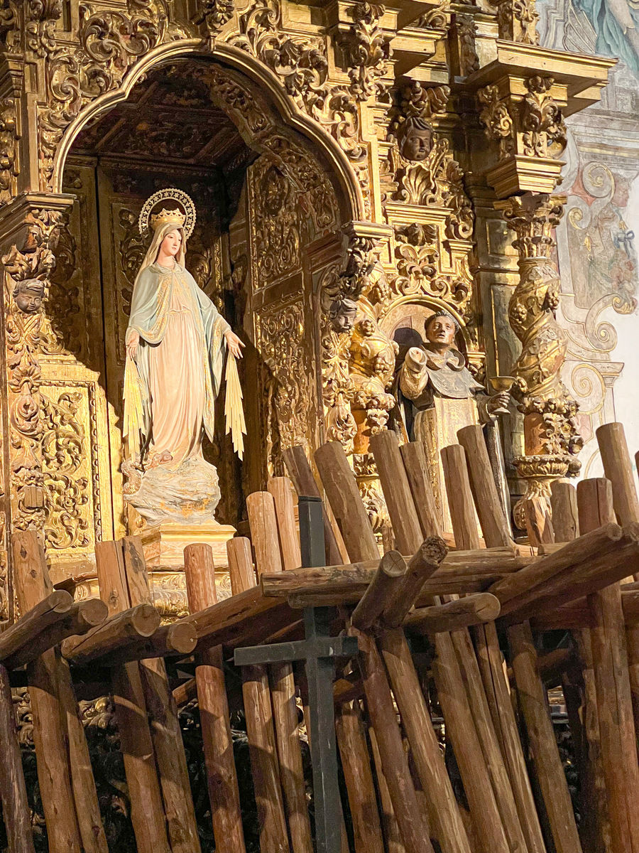 Posąg Maryi w kościele w Sewilli zdobionej, złotej kaplicy. Na pierwszym planie drewniane krzyże zgromadzone po procesji. Scena religijna, pełna spokoju i majestatu.