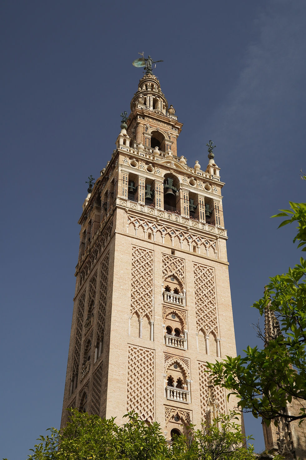 Wieża Giralda w Sewilli, z ceglaną ornamentyką, u góry figura, na tle błękitnego nieba. Na dole zielone liście, architektura gotycko-mudejarska.