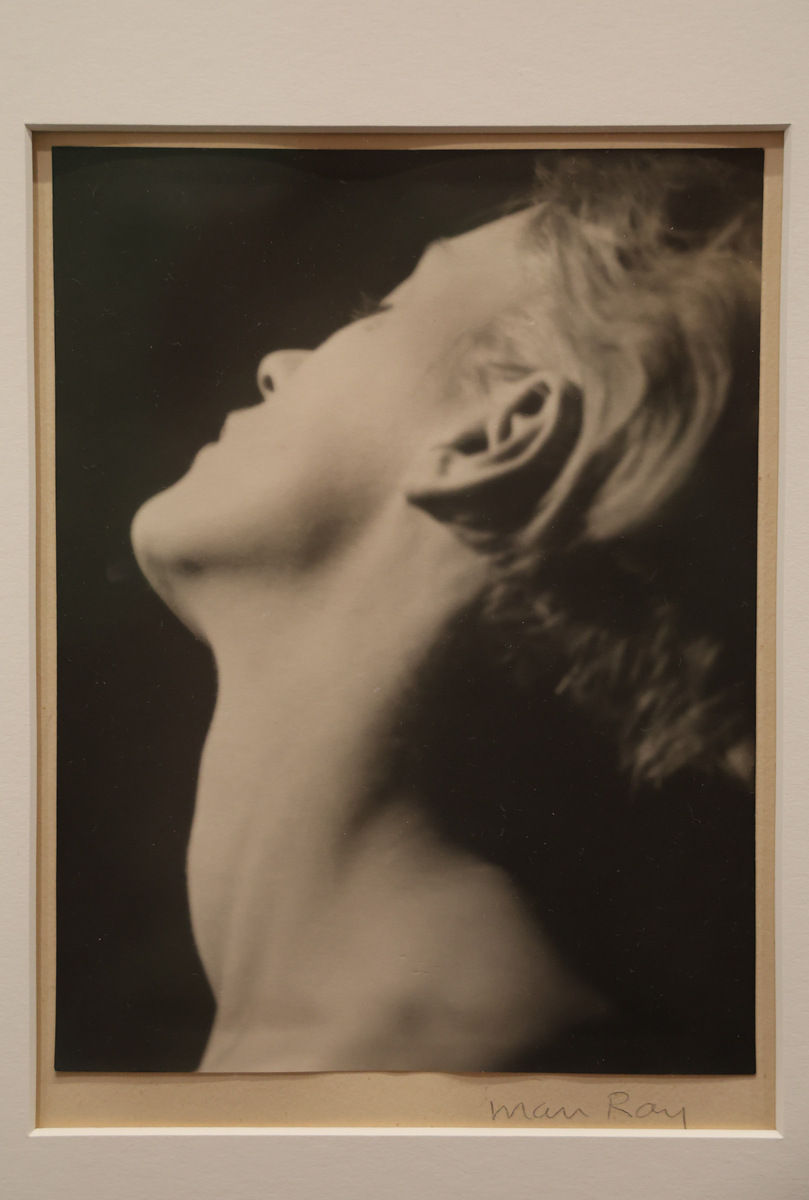 Czarno-biały portret kobiety z odchyloną głową w profilu; na dole podpis Man Ray.