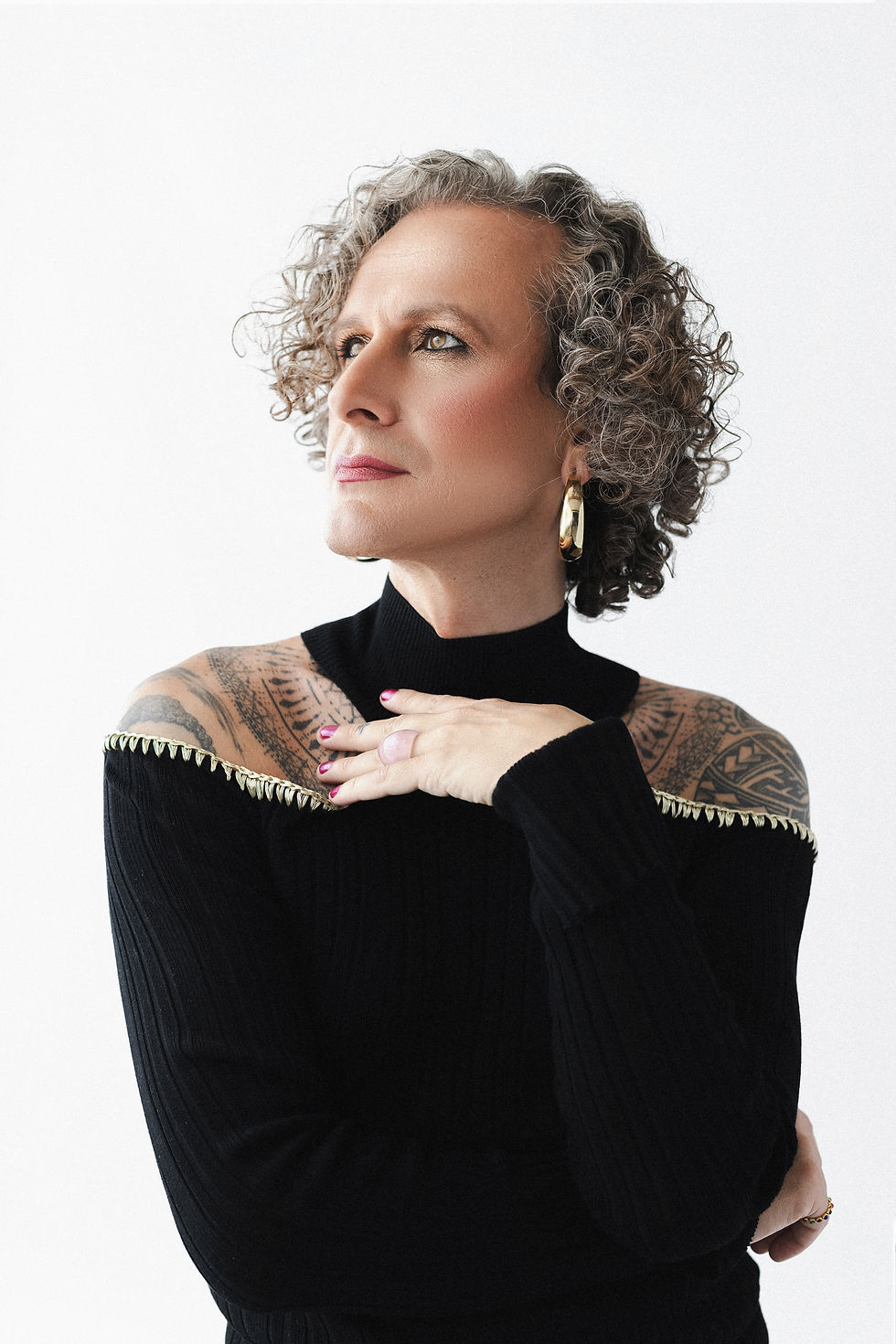 Leia Vallante — editorial trans model with shoulder tattoos in black high-fashion top, gazing boldly into the light. Queer be