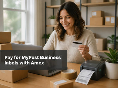Small business owner holding a credit card at a desk with a laptop, parcels and a shipping label printer.