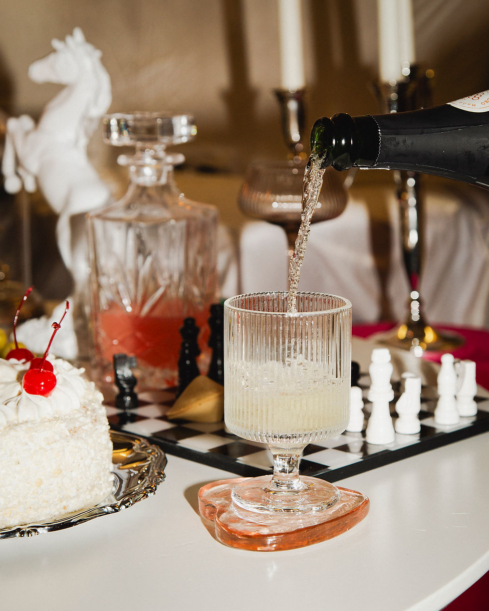 Kuohuviiniä kaadetaan kristallilasiin, taustalla shakkilauta ja kermakakku, jonka päällä punaisia kirsikoita.