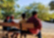 Four older adults sit in a row on a wooden bench facing a sand dune, green leaves of some large oak trees in the background catch the sunlight.