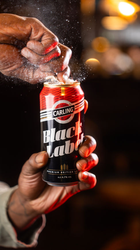 Two hands holding a Carling black label can. One is at the base while the other one is cracking the tab to open it, there is a spray of beer coming out of the can. The lighting feels bright on the can. Taken in Bristol Bar The Milk Thistle 