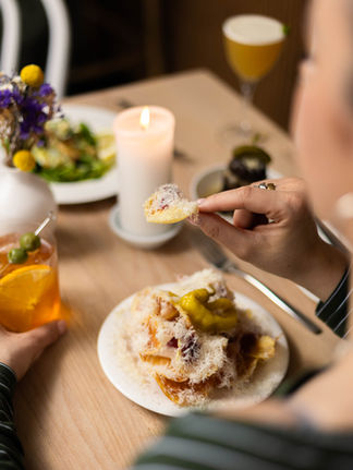 A women sat at a restaurant table holding a drink with food in the other hand. The table is a light wood with a tall candle in the middle. There is another dish and cocktail on the other side of the table.