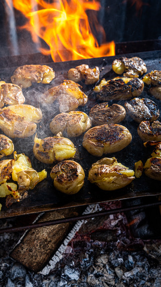 A tray of lots of potatoes is say on top of a wood fire. In the background there is corrugated iron and a large orange flame. 