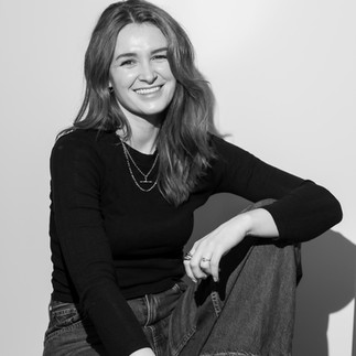 A black and white image of a women sat with one leg pull up towards her chest. She is wearing dark trousers and a long sleeve black top. She is smilling looking at the camera. Her hair has a loose wave to it. Shot in Future Proof photography studio