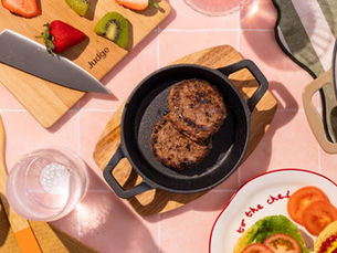 A flat lay image of a tiles table. The surface is pink times on the tiles here are judge cookware pots, pans and chopping boards. inside and on the cookware is food this includes chilli, chicken wings, burgers and sweetcorn.