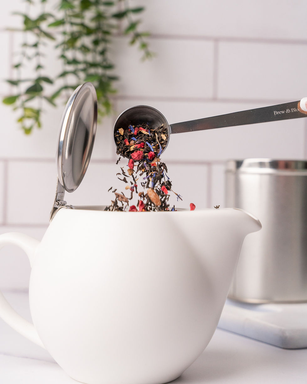 A white tea pot is sat on a white kitchen counter. There is a green plan coming down in the background on the left while a metal tin is on the right. A spoon is tipping a loose leaf tea into the tea pot. For tea company Brew and Bloom shot in Bristol located studio Future Proof.