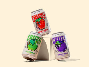 On a cream background there is a cream plinth with three drink cruzy can leaned against it. The lighting is strong casting harsh shadows on the drink cans.