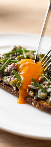An up close shot of a signiture Cor dish. On a white plate there is a slice of sourdouch with beef tar tar on shop garshied with an egg yolk which is being split with a knife and fork and the yolk is running down the bread.
