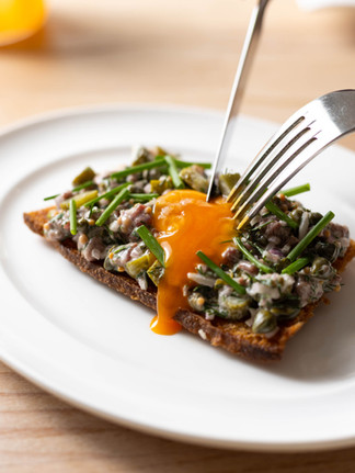 An up close shot of a signiture Cor dish. On a white plate there is a slice of sourdouch with beef tar tar on shop garshied with an egg yolk which is being split with a knife and fork and the yolk is running down the bread.