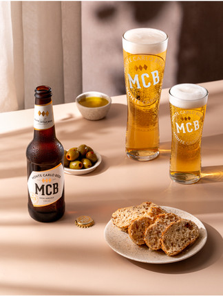 On a cream table surface there is an open bottle of monte carlo beer to the left to the right there is a pint and half pint of beer. In the fourground there is some slices of bread on a cream plate in the centre there is a small bowl of olives and olive oil. The background is a soft curtin. The lighting is soft and casting a dappled shadow effect.