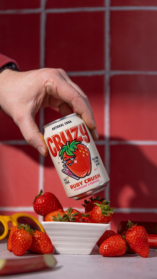 A hand is holding a can of ruby crush fizzy drink on an angle beneath there is a small pot of strawberry and to the left id a pair of yellow sunglasses. The background is a red tile and the surface is a grey concrete.