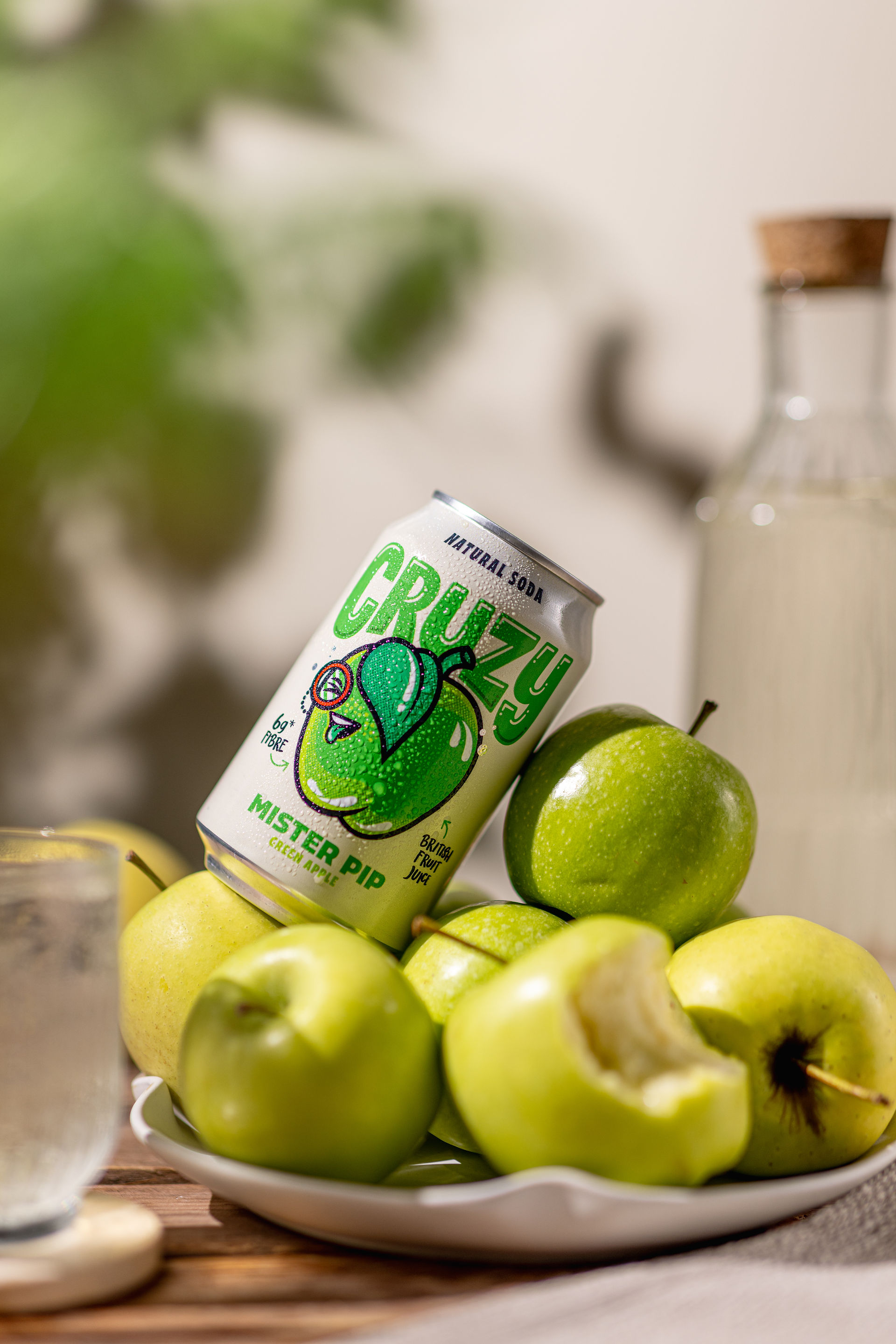 In the centre of the frame there is a plate with a large pile of apples on it. A can of mister pip is leaning against the apples. To the left is a glass of clear liquid. In the foreground there is a few leaves which are also casting a dappled shadow on the background which is cream. The surface is wooden boards. 
