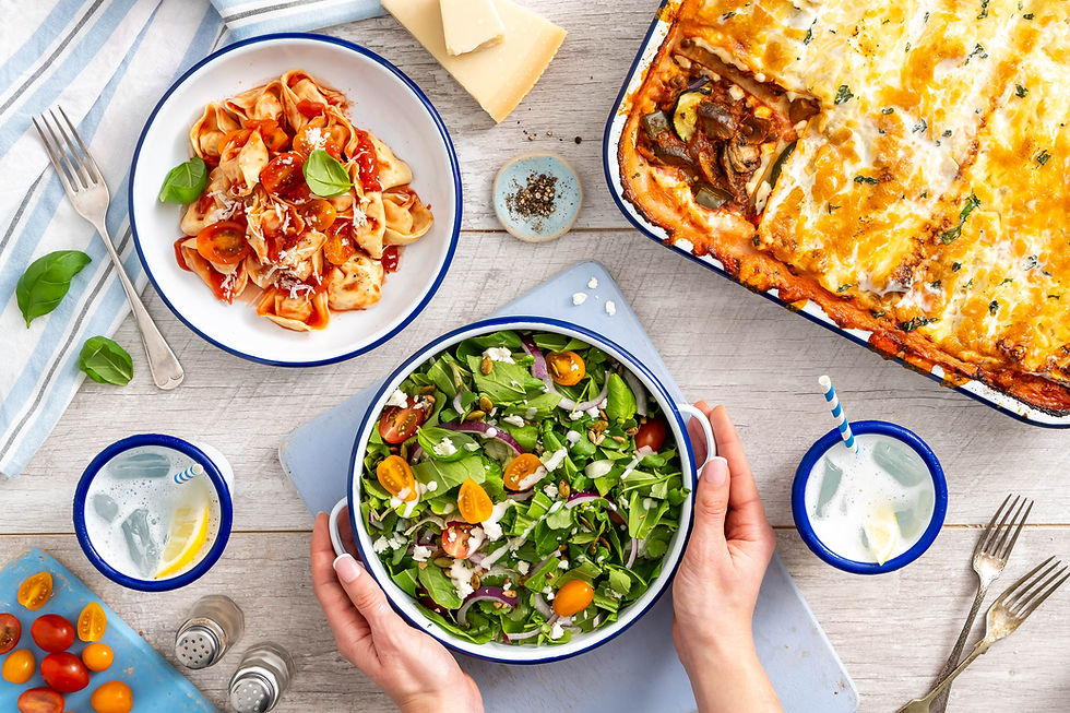 A grey wooden table scape with multiple dishes. On the left there is a stripped napkin with a Falcone enamelware bowl on it. In the bowl is a filled pasta in a tomato sauce topped with basil and cheese. In the middle there is a larger dish with a person holding it on either side. A green salad topped with fresh tomatoes and feta is in the dish. To the right is a much larger dish with a lasagne in it. This was taken in Bristol photography studio Future Proof. 