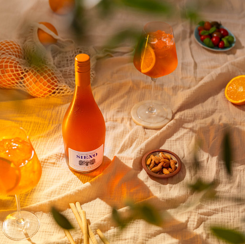 A warm picnic set up inclued a lines background as the base. In the shot these is a mixtures of oranges in a bag, a selection of nuts and tomatoes. In two wine glasses there is orange liquid. In the centre of the frame there is a bottle of non alcoholic orange spritz. Shot in Bristol photography studio   