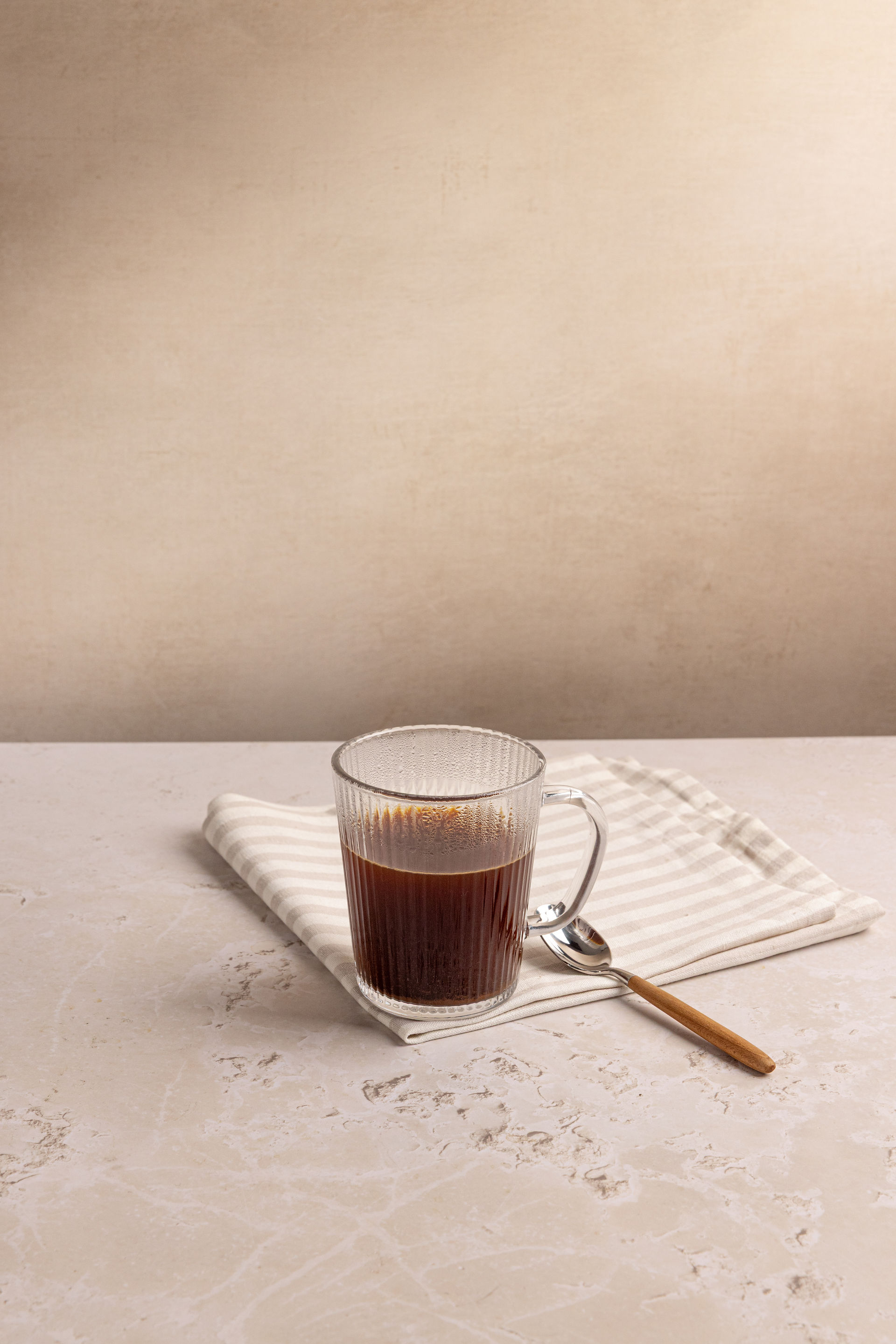 A glass mug is in the centre of the frame on a stripy tea towel with a spoon to the right hand side. The back ground is cream and the surface is  a textured cream. Shot in Bristol based photography studio Future Proof.