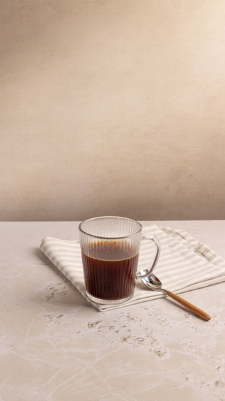 A glass mug is in the centre of the frame on a stripy tea towel with a spoon to the right hand side. The back ground is cream and the surface is  a textured cream. Shot in Bristol based photography studio Future Proof.