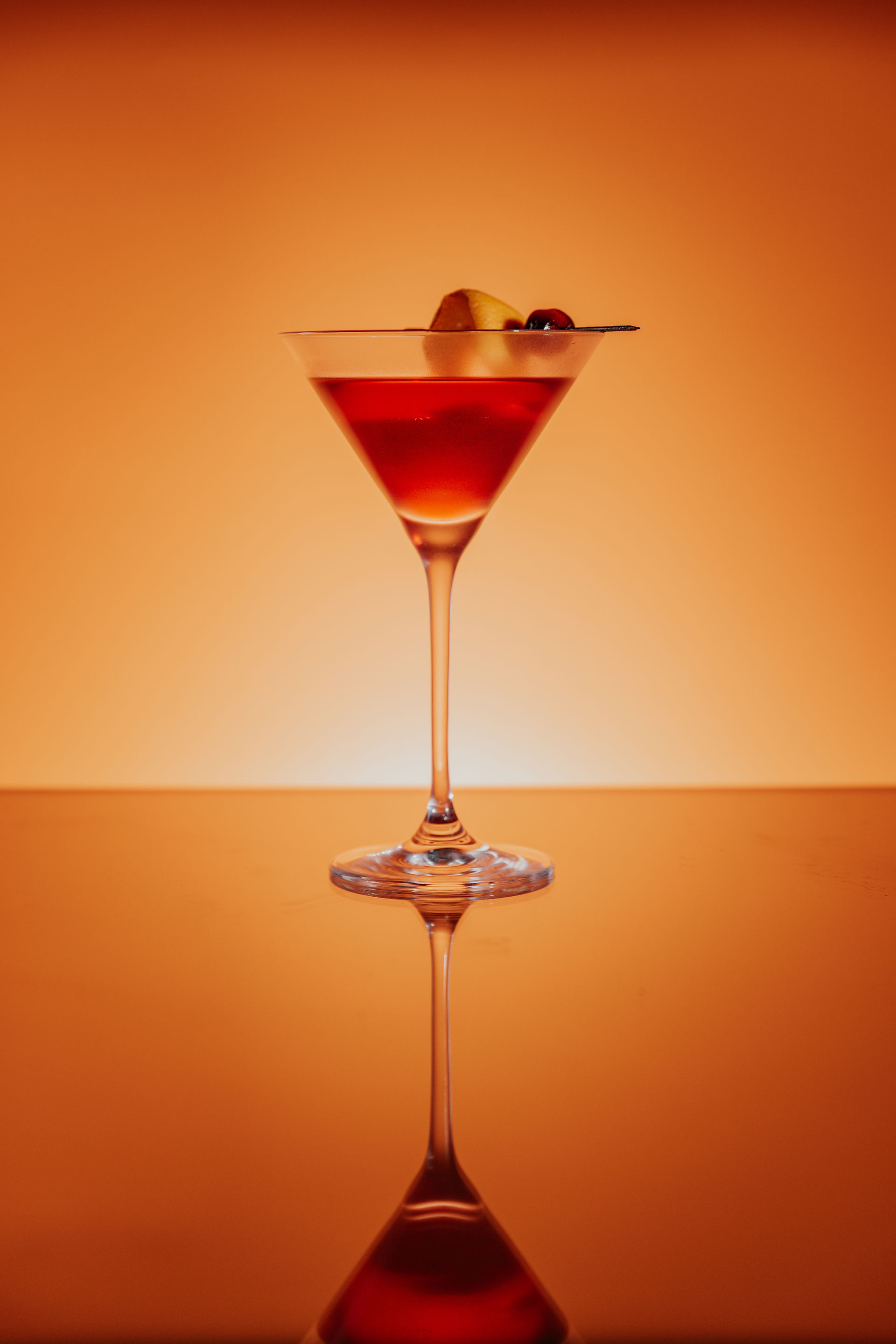 Drink photography of a red cocktail in a martini glass. The surface is reflecting the glass and the background is orange with light glowing from below.