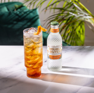 Shot in Bristol based photography studio a glass and bottle of double dutch are in the middle of the frame on a white marble table. In the background there is a large plant and green velvet chair. The lighting is casting harsh shadows.