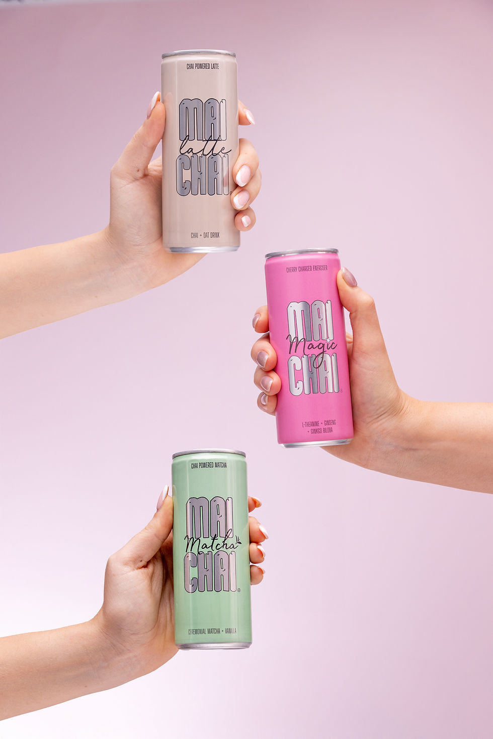 Three cans of Mai Chai are held up against a soft pink background. Two hands are coming in from the left and one is coming in from the right. The lighting is soft highlighting the logo on the can. Shoot at Future Proof Photography studio Bristol.