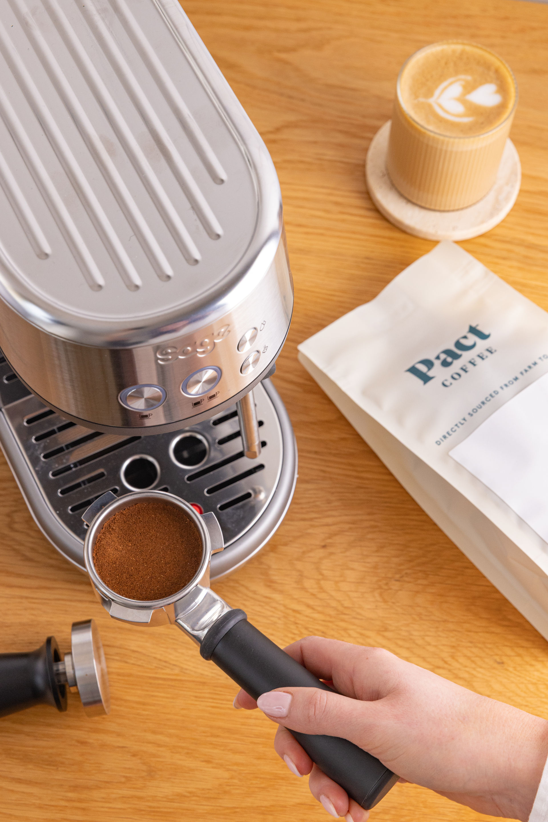 On a wooden surface there is a latte with some heart latte art on it to the right hand side. Under this is a bag of pact coffe. To the left of the shot is a silver ocffee machine with a hand coming in from the bottom the the frame holding the handel of the coffee basket as if its about to be placed in to brew.
