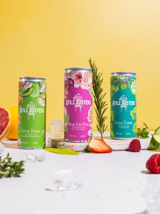 Three cans of Still Sister gin are in the centre of the frame with flavour ques including rosemary, strawberries and cucumber. The surface is white and the backdrop is yellow. Shot in Bristol studio Future Proof.