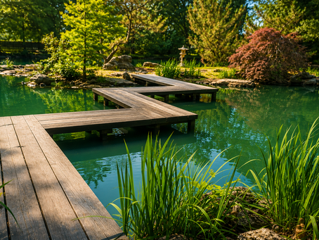 A peaceful view of the Mizumoto Japanese Stroll Garden in Springfield, Missouri. A zigzagging wooden boardwalk stretches across tranquil jade-green water toward a small island adorned with trees, natural stone, and a traditional Japanese stone lantern. Sunlight filters through the surrounding evergreens and Japanese maples, casting warm reflections across the pond and creating an atmosphere of stillness and contemplation. This photo was taken by the author and has been enhanced using AI to improve composition, lighting, and color while preserving the spirit of the original photograph.