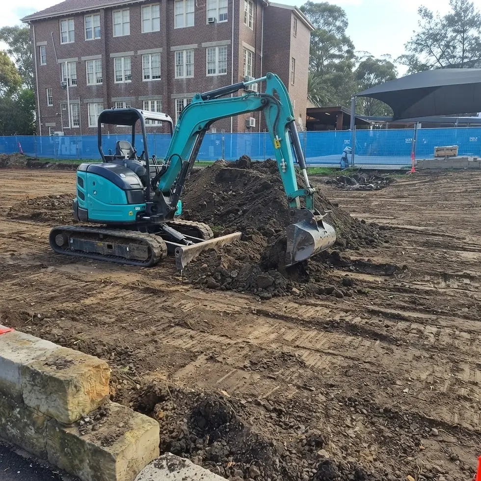 Outdoor photography of a小型 excavator digging into a pile of soil on a construction site. The image captures a routine operational scene with a multi-story building in the background.