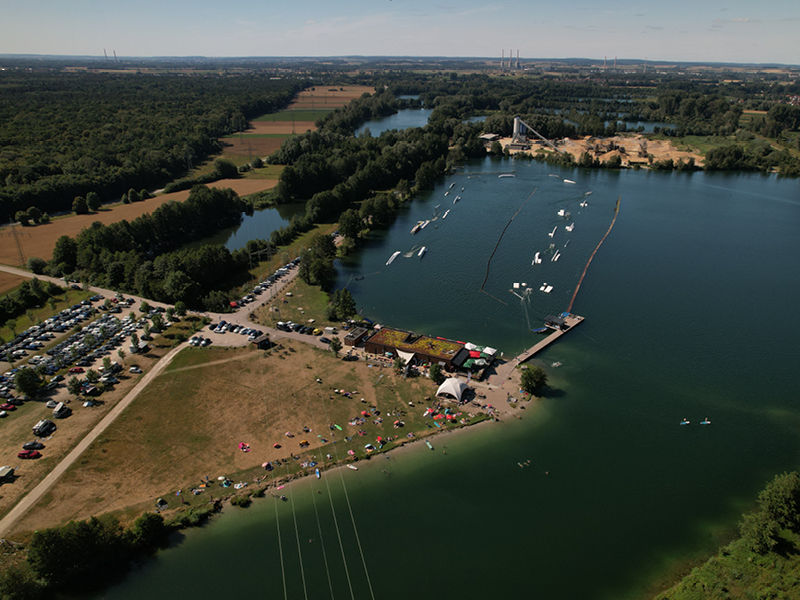 Luftaufnahme des Wakeparks von Wake & Groove mit See, Cable-Anlage, Liegewiese und Stellplätzen am Ufer.