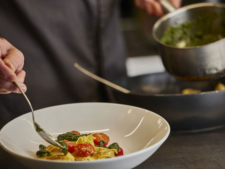 Pasta wird in einer Küche angerichtet, mit grüner Sauce und Tomaten in einem weißen Teller.