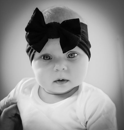 Baby girl in a white vest and black bow on her head, sat upright looking directly at the camera.