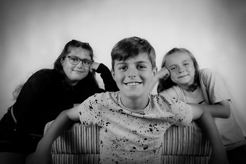 Family picture of three children. The brother is sat at the front and both his sisters are behind him on both sides. all smiling at the camera. White background