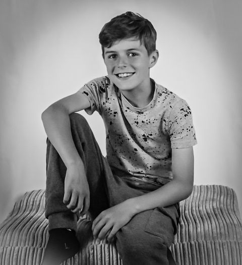 Young child, sat on the edge of a chair with one leg bent and smiling into the camera. White background and grey clothing, A very relaxed pose