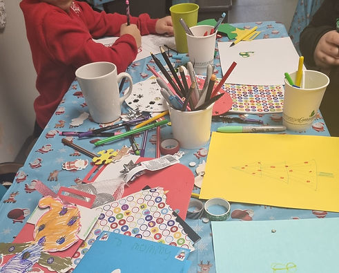 A tbale full of arts and crafts with a child form the neck down colouring a picture.