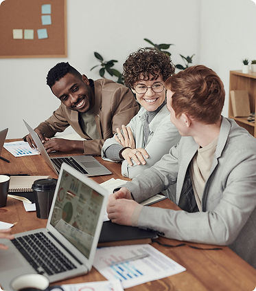 reunião de trabalho com 3 colaboradores conversando, dois sorrindo, em uma mesa de escritório com computadores e papeis