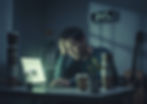 Man at desk, hand on head, looking tired at laptop. Clock shows 3:47 AM. Stacks of coffee cups, guitar in background, dim lighting.