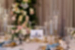 Elegant table setting with pink and white flowers, tall candles in glass holders, gold accents, and a "Head Table" card against a floral backdrop.