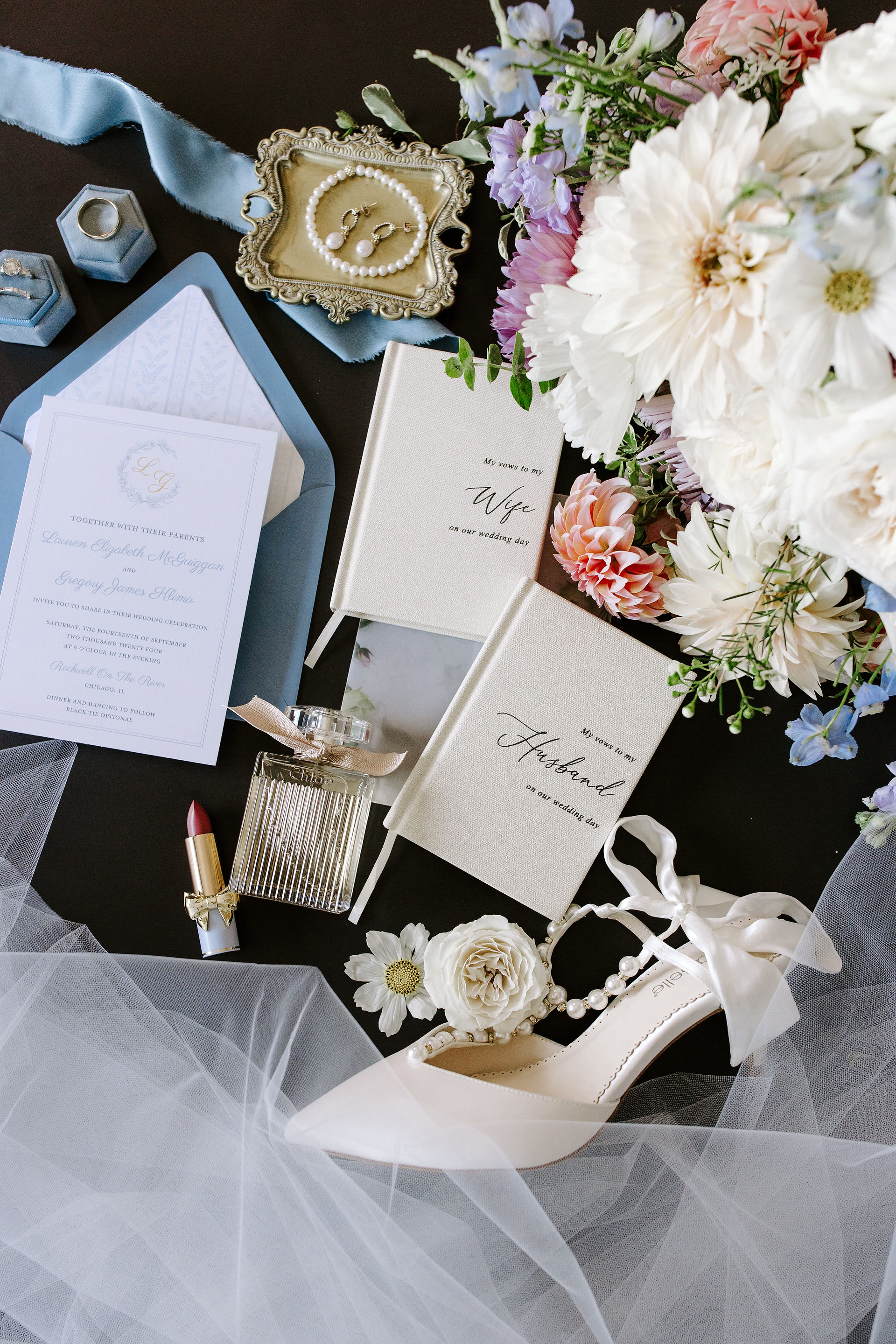 A styled wedding flat lay featuring an open invitation, white high-heeled shoe, pen, and floral arrangement on a dark background.