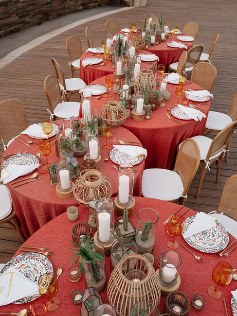 Alt text Aerial view of multiple bridal shower tables styled with terracotta linens and botanical dinnerware on an oceanside terrace, with rattan lanterns and pillar candles set against a coastal stone backdrop.
