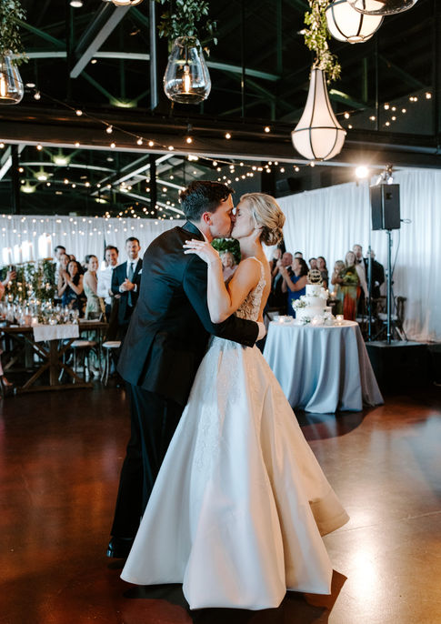 Bride and groom sharing their first kiss on the dance floor at Theater on the Lake Chicago, surrounded by applauding guests, with pendant lanterns, string lights, and tall greenery centerpieces illuminating the reception space behind them.
